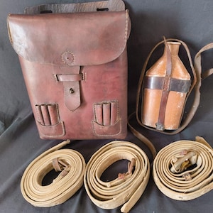 May include: A collection of vintage leather items, including a brown leather satchel with two front pockets, a canteen with a strap, and three rolled canvas straps. The satchel has a flap closure and a button detail.