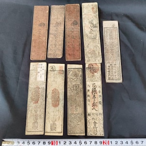 May include: A collection of antique rectangular paper documents with faded colors and intricate text and designs. The papers vary in shades of brown and beige, with some featuring red seals. The documents are arranged on a dark surface.