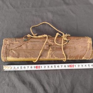 May include: A worn, brown leather case with a drawstring closure. The case has a rectangular shape with a pocket on the front. A measuring tape is placed next to the case.