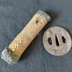 Op de afbeelding: Een cilindervormig zwaardhandvat omwikkeld met getextureerde, crèmekleurige roggenhuid, voorzien van een donkere metalen dop aan de bovenkant en een ruwe, grijze basis, naast een ronde, verweerde metalen stootplaat met reliëf.