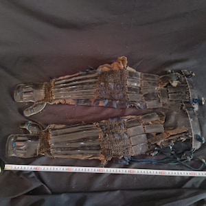 Puede incluir: Par de antiguos guanteletes de samurái japonés. Los guanteletes son negros con placas articuladas, conectadas por cota de malla. Los guantes tienen un forro azul oscuro y se exhiben sobre una superficie negra. Los guanteletes miden aproximadamente 30 cm de largo.