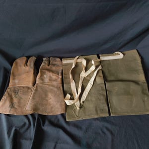 May include: A set of vintage work gloves and a canvas apron. The gloves are brown leather with a split-toe design. The apron is olive green canvas with cream-colored straps. The gloves have faded markings.