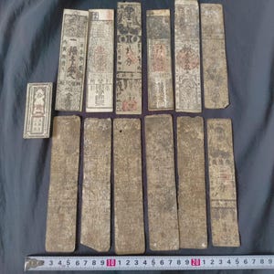 May include: A collection of thirteen rectangular paper slips featuring aged, off-white surfaces with black ink calligraphy, red stamps, and intricate illustrations. A metal measuring tape at the bottom shows the items range up to 9 inches in length.