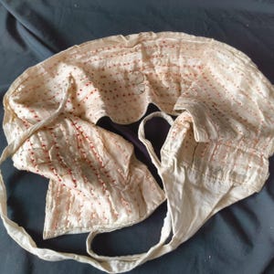 May include: Vintage cream-coloured bonnet with red stitching and a button fastening. The bonnet features a scalloped edge and long ties. The fabric appears to be a soft material, possibly silk. The bonnet is likely from the Victorian or Edwardian era.