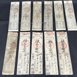 May include: A collection of antique Japanese paper money, featuring intricate designs and calligraphy. The rectangular notes display red seals and detailed text, with varying shades of gray and brown. The notes are arranged on a dark surface.