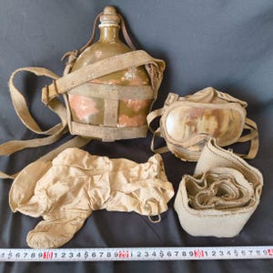 May include: An assortment of vintage military gear, including a green canteen with a canvas strap, goggles, gloves, and a rolled-up cloth. The items are in various shades of tan and olive green, suggesting age and use. The items are arranged on a dark surface.