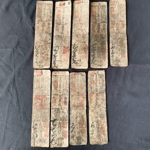 May include: A collection of ten rectangular, aged paper currency notes. The notes are primarily beige with red and black ink markings, including text and seals. The paper shows signs of wear and age, with visible creases and discoloration.