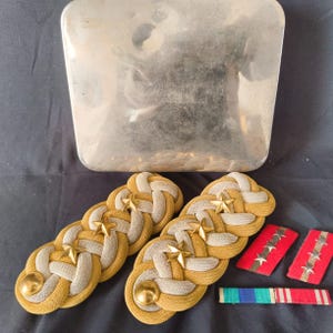 May include: Military insignia set featuring two pairs of gold and grey shoulder boards with star details, two red rank badges with stars, and a ribbon bar. A clear, rectangular object is in the background.