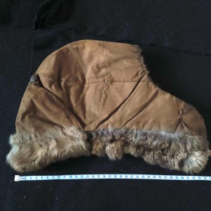 Op de afbeelding: Een bruine wintermuts met een bontrand langs de onderrand. De hoed is gemaakt van een zacht, suède-achtig materiaal en heeft een capuchon. De bontrand is een mix van bruin en grijs. De hoed ligt op een zwart oppervlak.