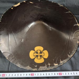 Pode incluir: Um chapéu japonês antigo, preto e redondo, com um emblema dourado. O chapéu tem uma forma ligeiramente cónica e mostra sinais de idade, com rachaduras e desgaste nas bordas. Uma régua é colocada por baixo.