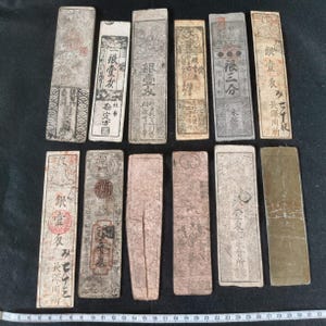 May include: A collection of antique Japanese paper money, featuring various rectangular denominations with intricate designs and Japanese characters. The aged paper has a faded appearance, with visible wear and tear. Some pieces have red seals and stamps.