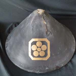 Pode incluir: Um chapéu cinza escuro em forma de cone com um emblema dourado com cinco círculos dentro de um quadrado. O chapéu mostra sinais de desgaste, com alguns danos visíveis. O design do chapéu sugere um significado histórico ou cultural.