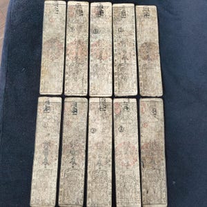 May include: A collection of ten rectangular, aged paper cards. Each card features intricate, faded illustrations and text in a traditional East Asian style, with red circular seals. The cards are arranged on a dark blue surface.