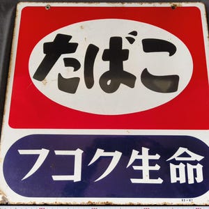 Könnte beinhalten: Vintage japanisches Metallschild mit schwarzer japanischer Schrift in einem weißen Oval auf rotem Grund. Darunter befindet sich ein blaues Oval mit weißer Schrift. Das Schild weist Gebrauchsspuren auf.