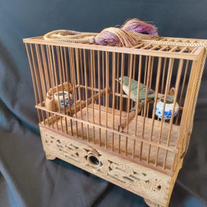 Puede incluir: Jaula de pájaros de madera clara hecha a mano con un pequeño pájaro verde y azul en su interior. La jaula presenta intrincadas tallas en la base, barras verticales y un detalle decorativo de cuerda y borla en la parte superior. Dos pequeños cuencos de cerámica en el interior.
