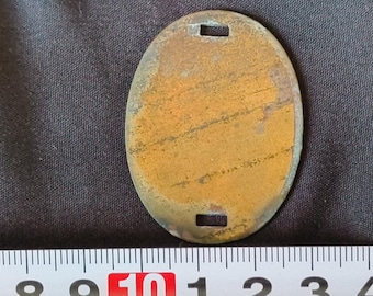 May include: A brass oval-shaped tag with two rectangular holes at the top and bottom. The tag is slightly tarnished and has a worn, aged appearance.