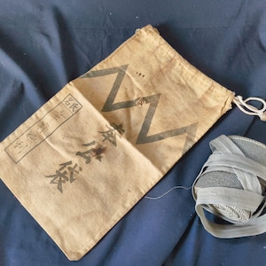 May include: Tan drawstring bag with Japanese characters and a zigzag design in black. A bundle of gray ribbon is next to the bag. The bag has a white drawstring and is on a dark blue surface.