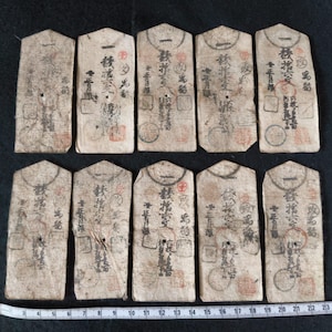 May include: Ten antique Japanese paper currency notes. Each note is rectangular with a pointed top and features intricate calligraphy and red seals. A ruler with inch measurements is at the bottom.