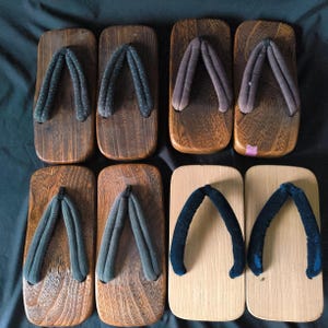 May include: An overhead shot of eight pairs of geta, traditional Japanese wooden sandals. The wooden platforms vary in color from light to dark brown. The thong straps are in shades of blue, gray, and brown. The sandals are arranged in two rows of four.