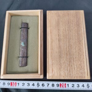May include: A rectangular, dark brown metal object with etched designs, held in a wooden box with a green interior. The box is open, revealing the object, and the lid is beside it. The object is secured with two black bands.