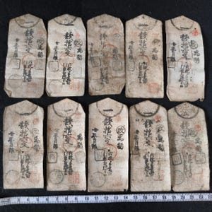 May include: A collection of ten aged, rectangular paper items with Japanese calligraphy and red seals. The paper has a worn, textured appearance with visible creases and stains. The text is in black ink, and the overall aesthetic suggests an antique or historical artifact.