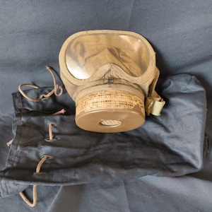 May include: An olive-drab gas mask with clear lenses and a circular filter. The mask has a fabric body and adjustable straps. A black fabric bag with drawstrings and eyelets is also present. The mask has Japanese text on the filter.