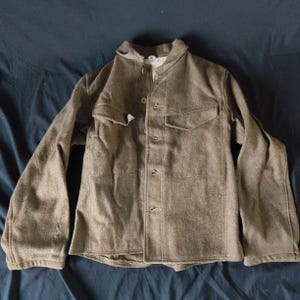 May include: A vintage olive-drab wool jacket with a button-up front, two chest pockets, and a collar. The jacket has long sleeves and appears to be a military-style garment. The fabric has a textured appearance.