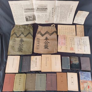 May include: A collection of vintage Japanese documents and books. The image shows two drawstring bags with Japanese characters, alongside various papers and small books of different colours and sizes. The items are arranged on a dark blue surface.