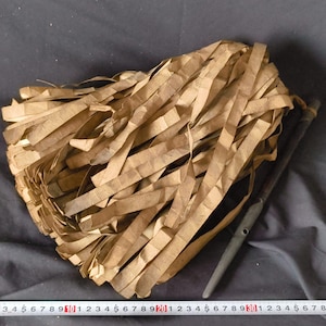 May include: A bundle of brown paper strips, tightly packed together, with a dark, slender object alongside. The paper appears to be aged or vintage, with a slightly textured surface. The dark object has a pointed end and a hole.