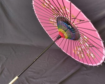 May include: Pink Japanese parasol with a black handle and ribs. The parasol features floral designs and a colorful interior. The handle has a white tip. The parasol is open, displaying its full circular shape.