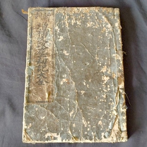 May include: An antique book with a weathered, slate-gray paper cover featuring visible creases, worn edges, and a rectangular label on the left side containing vertical black Japanese text. The binding is secured with visible thread stitching.