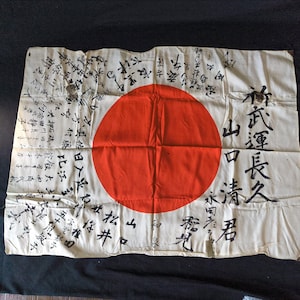 Puede incluir: Bandera japonesa vintage con un gran círculo rojo en el centro, que representa el sol. La bandera es de color blanquecino con inscripciones caligráficas negras. La bandera rectangular parece ser de tela.