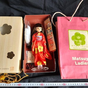 May include: A Japanese doll in a red kimono with yellow floral accents, presented in a wooden box and a pink shopping bag. The doll is approximately 6 inches tall. The box and bag feature the "Matsuya Ladies" logo.