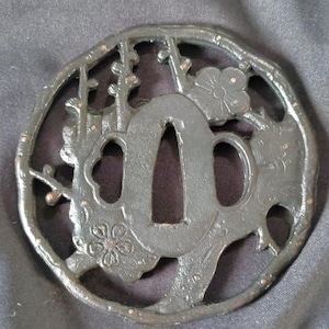 Op de afbeelding: Een cirkelvormige tsuba van donkergrijs metaal met een opengewerkt ontwerp van pruimenbloesemtakken en bloemen. Het midden heeft een langwerpige ovale opening voor een zwaardblad, geflankeerd door twee kleinere openingen, alles in getextureerd metaal.