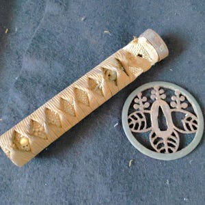 May include: A cylindrical sword handle wrapped in light tan cord over a textured cream-coloured ray skin base, accompanied by a circular metal hand guard featuring an intricate openwork design of stylised leaves and a central oval opening.