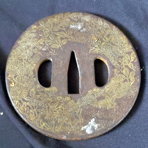 Op de afbeelding: Een cirkelvormige metalen tsuba zwaardbeschermer met een ingewikkeld gegraveerd bloemmotief met goudkleurige accenten. Het stuk heeft een centrale taps toelopende opening geflankeerd door twee kleinere ovale uitsparingen met een donkere patina.