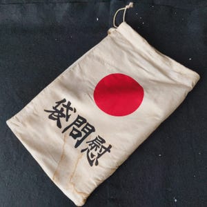 May include: Cream canvas bag with a red circle and Japanese characters in black. The bag has a drawstring closure and shows signs of age, with staining. The design is similar to the Japanese flag. The bag is likely used for storage.
