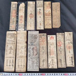 May include: A collection of antique paper currency, featuring various rectangular notes with intricate designs and Chinese characters. The notes display red seals and faded ink, suggesting age and historical significance. Some notes are folded or slightly damaged.