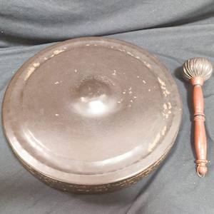 Può includere: Una ciotola tibetana color bronzo con un battente in legno abbinato. La ciotola è rotonda con un centro leggermente rialzato e un bordo decorativo. Il battente ha un manico in legno e una testa arrotondata strutturata.