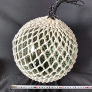 Könnte beinhalten: Ein runder, klarer Glas-Schwimmer, der in einem weißen, gewebten Netz eingeschlossen ist. Der Schwimmer hat einen schwarzen Seilgriff an der Oberseite. Ein Maßband befindet sich am unteren Bildrand.