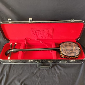 Może przedstawiać: Tradycyjne japońskie shamisen w futerale. Instrument ma długą, czarną szyjkę i okrągły korpus pokryty skórą węża. Futerł jest czarne z czerwonym wnętrzem.