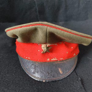 Könnte beinhalten: Vintage Militärmütze mit olivgrüner Krone und rotem Band. Die Mütze hat ein angelaufenes Metallstern-Emblem und ein rissiges, schwarzes Lederschild. Ein dünner roter Streifen säumt die Oberkante.