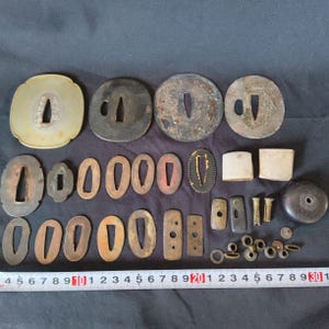 May include: A collection of antique Japanese sword fittings, including tsuba (guards), fuchi (collars), and other metal components. The pieces vary in shape, size, and color, with visible signs of age and wear. A ruler is placed at the bottom.