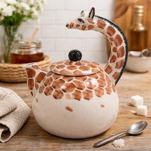 May include: A ceramic teapot featuring a giraffe design. The teapot is white with brown giraffe spots and a giraffe head handle. The lid has a black knob. A spoon and sugar cubes are on the wooden surface.