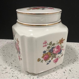May include: A white ceramic jar with a lid, decorated with pink roses, blue flowers, and green leaves. The jar has a gold trim. The jar has a unique, geometric shape. The jar is suitable for storing items.