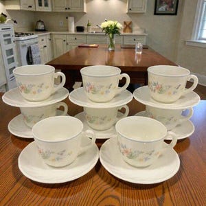 May include: A collection of six cream-coloured teacups and saucers. Each cup and saucer has a delicate floral pattern in pastel shades and a light blue rim. The cups are stacked on the saucers, arranged on a wooden table.