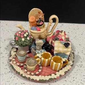 May include: A decorative figurine with a cat on a chair, a teapot, cups, spools of thread, and a floral arrangement. The base is a pink tray with a scalloped edge. The cups have yellow and white stripes.