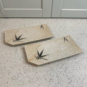May include: Two rectangular beige ceramic plates with a speckled texture. Each plate features a black bamboo leaf design. The plates are placed on a speckled white surface.