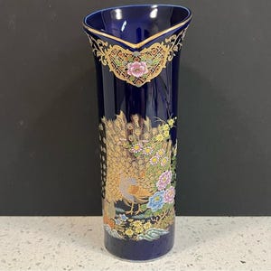May include: A tall, navy blue ceramic vase with a peacock and floral design. The vase has gold accents and a decorative rim. The peacock is shown with its feathers spread, and the flowers are in pink, blue, and yellow.