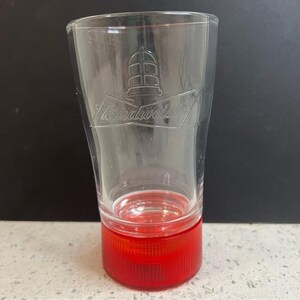 May include: A clear Budweiser beer glass with a red base. The glass has a tapered shape and the Budweiser logo is visible. The base is a solid red colour and has vertical ridges.